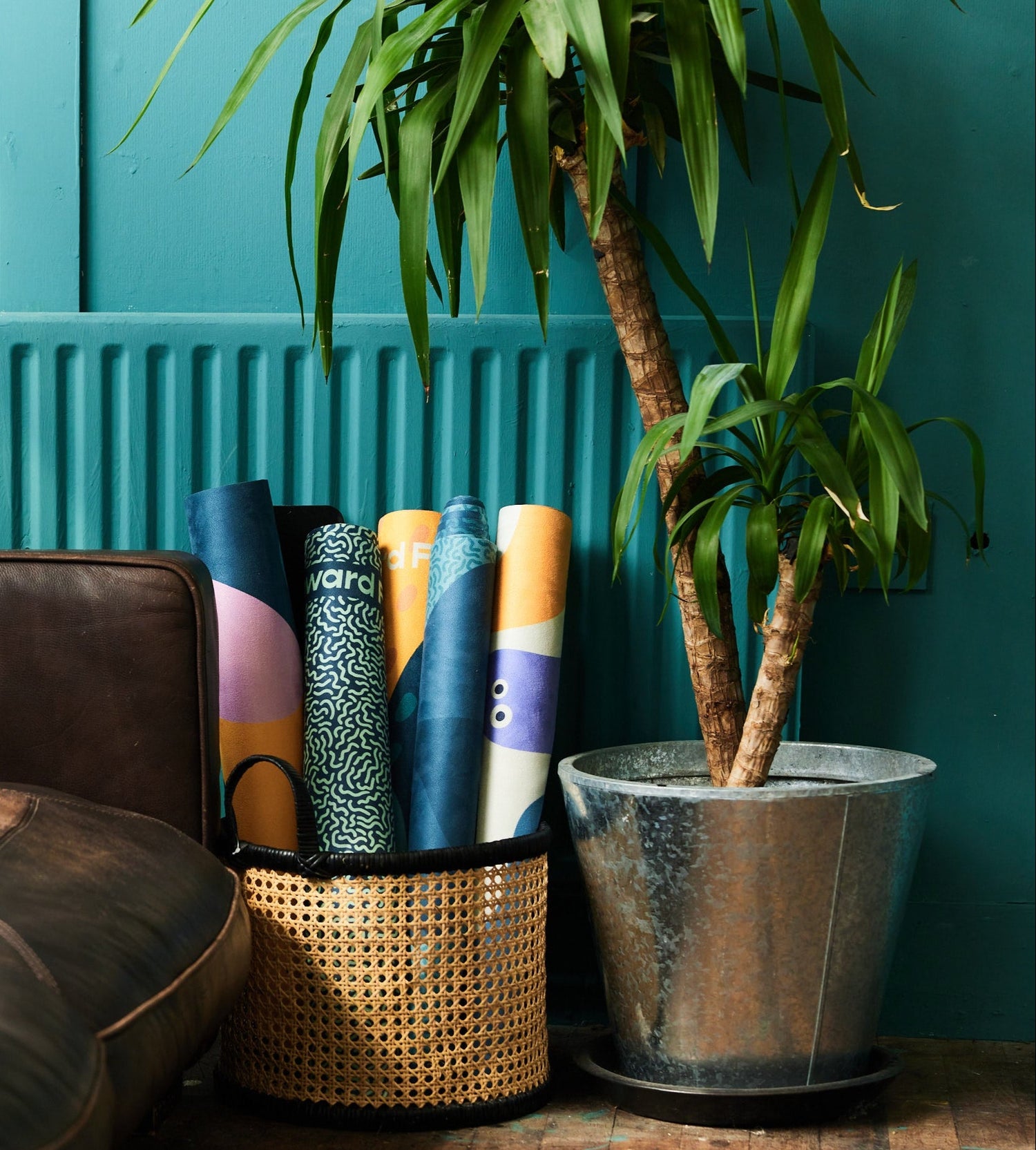 Brown leather sofa against a teal wall, featuring a wicker basket with rolled-up Downward Frog patterned yoga mats beautifully on display