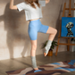 Woman in light blue shorts and gray top dancing in a room on top of a patterned yoga mat with plants and an easel.