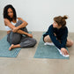 Two women sitting on yoga mats in a room with a white wall and wooden table.